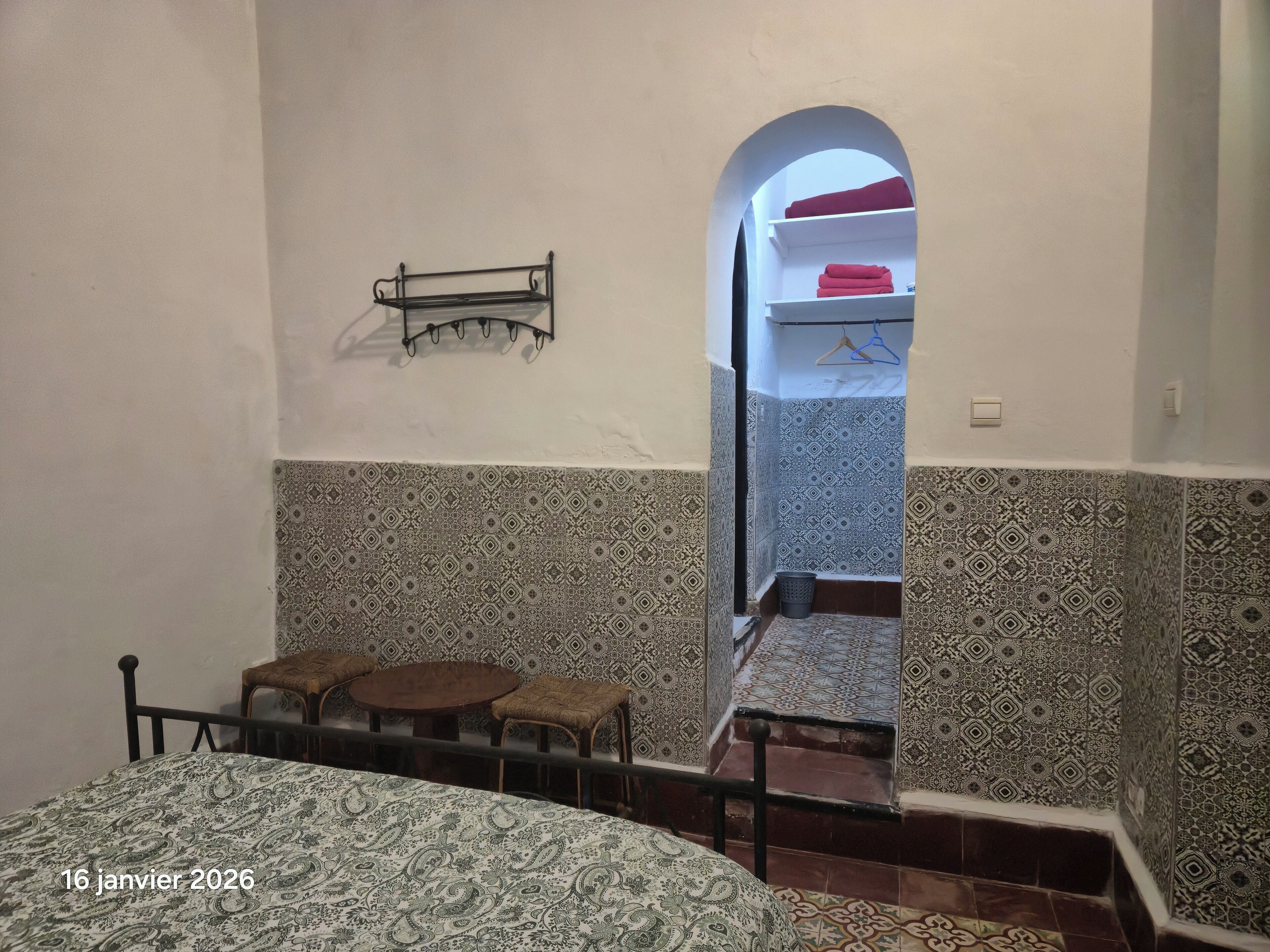 A bedroom with a bed, a small table, and a chair. There is a hanging rack on the wall and a staircase leading to an upper level.