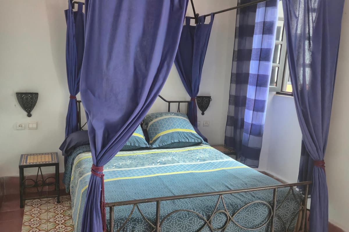 A bed with a canopy, a small table, a wall-mounted light, and a window with curtains.