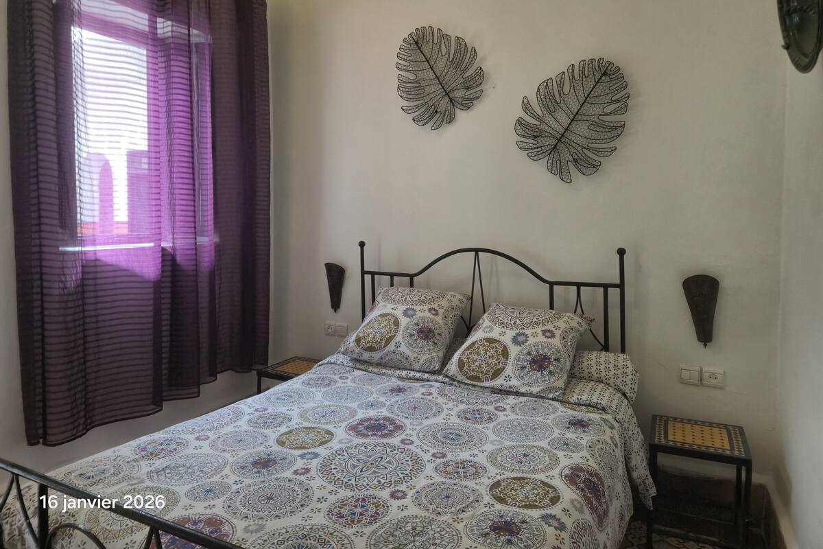 A bedroom with a bed, patterned bedding, a window with curtains, wall-mounted lamps, and leaf-shaped wall art.