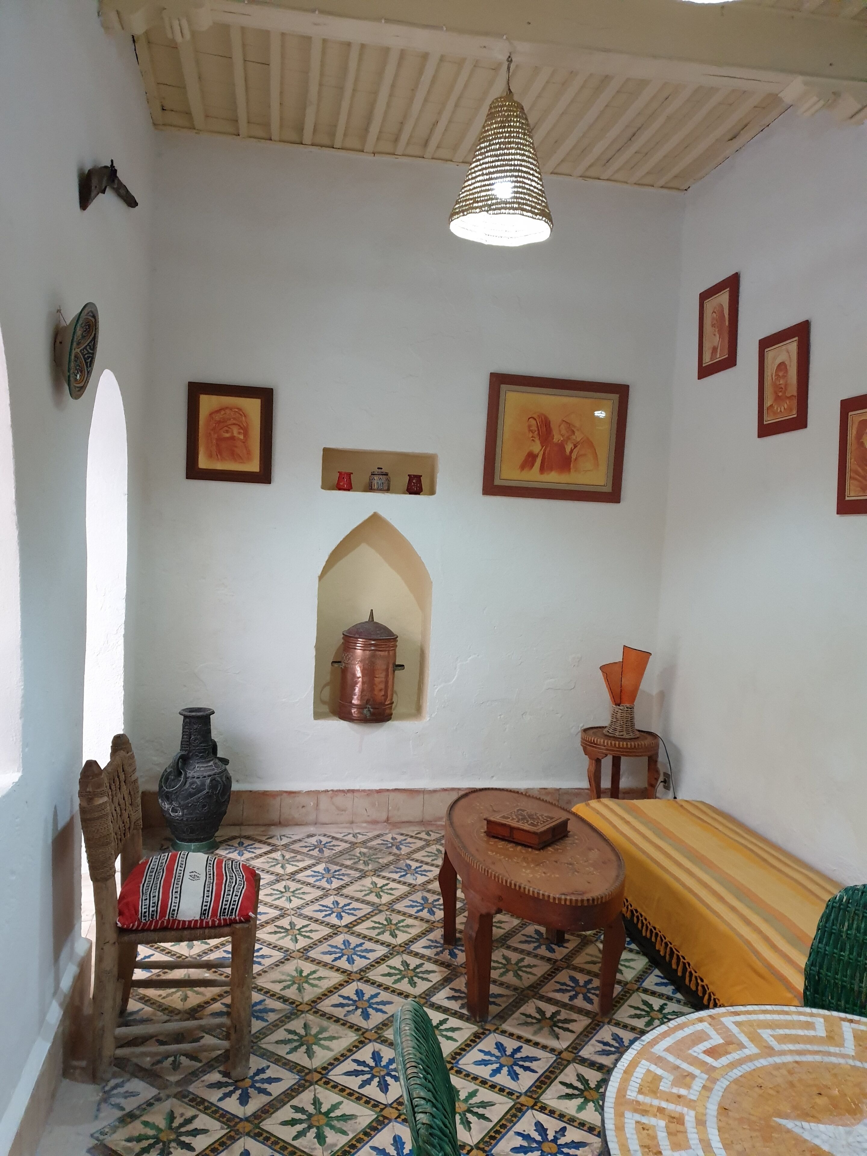 Una habitación con piso de baldosas estampadas, una silla de madera, una mesita pequeña, una lámpara colgante y varias fotografías enmarcadas en la pared.
