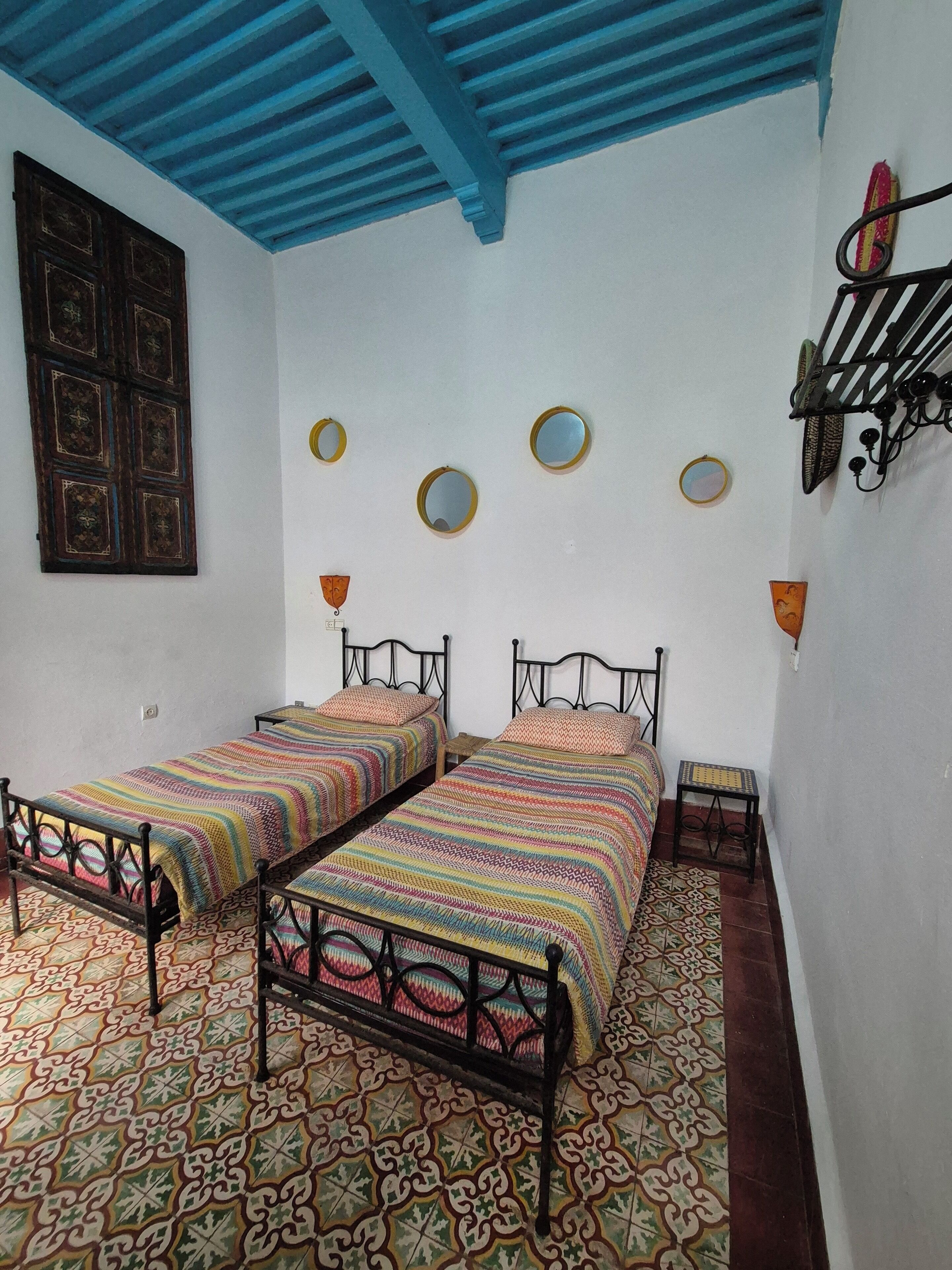 Une chambre avec deux lits, des carreaux de sol à motifs, un plafond bleu et des miroirs fixés au mur.