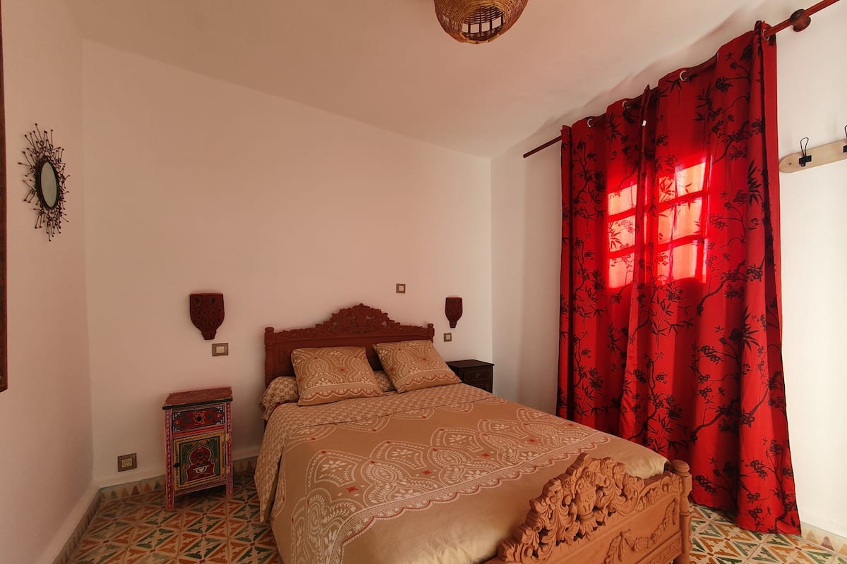 A bedroom with a wooden bed, patterned bedspread, red curtains, a decorative wall hanging, and a tiled floor.