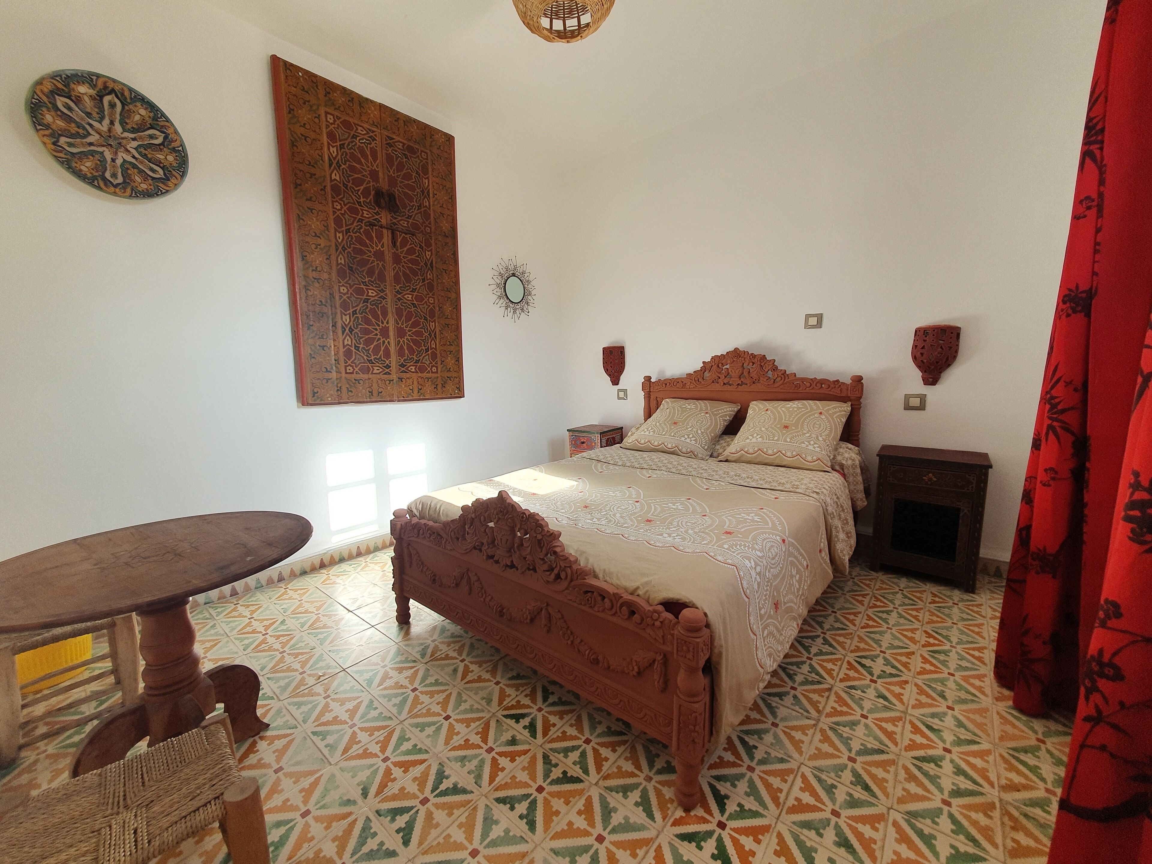 A bedroom with a bed, a small table, a decorative wall hanging, and a patterned floor.
