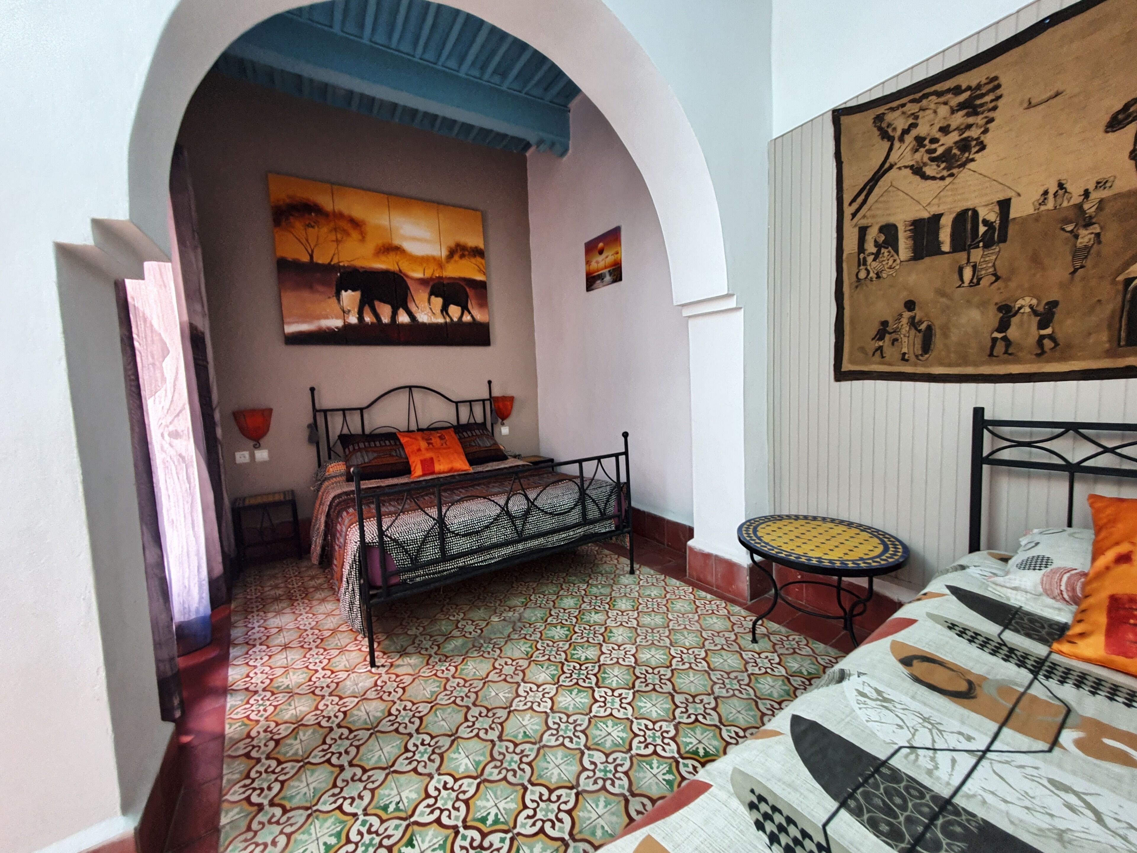 Une chambre avec un lit, une petite table, un mur décoré d’œuvres d’art et un sol recouvert de carreaux à motifs.