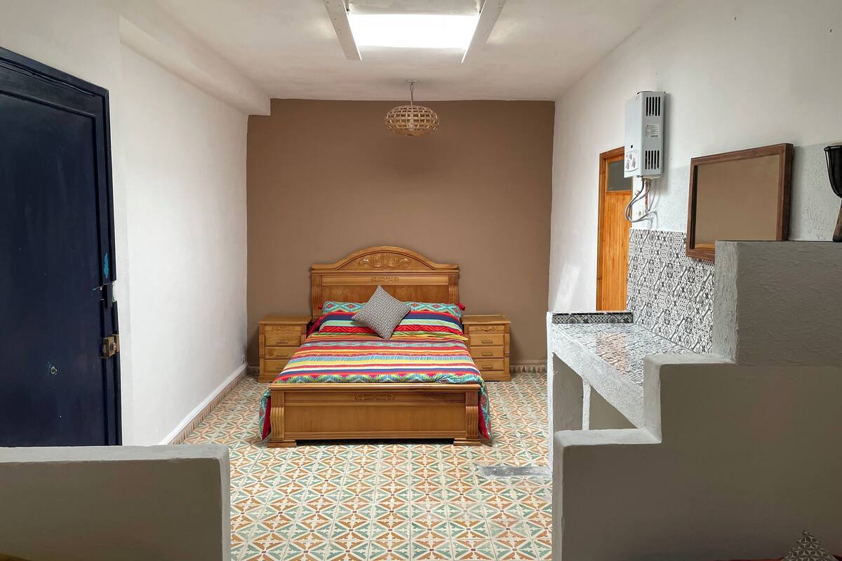 A bedroom with a wooden bed, a nightstand, a patterned floor, a ceiling light, and a wall-mounted air conditioner.