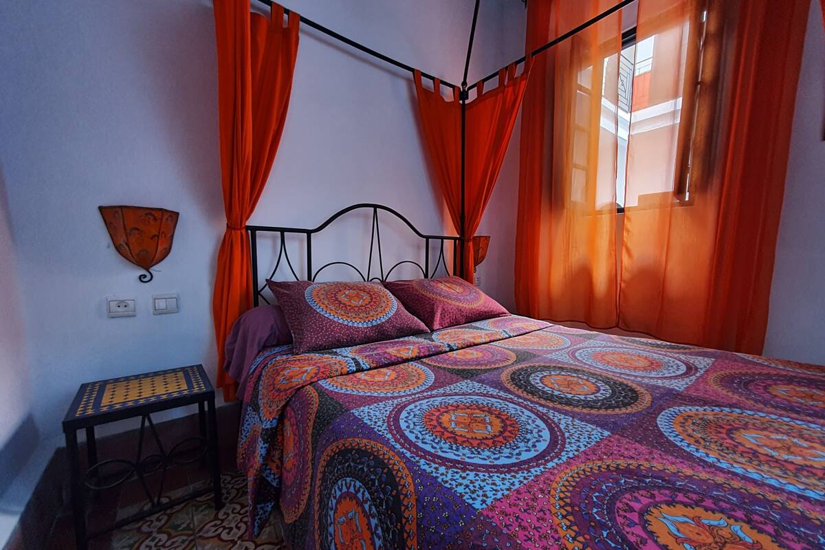 A bedroom with a bed, orange curtains, a patterned bedspread, a small table, and a wall-mounted lamp.