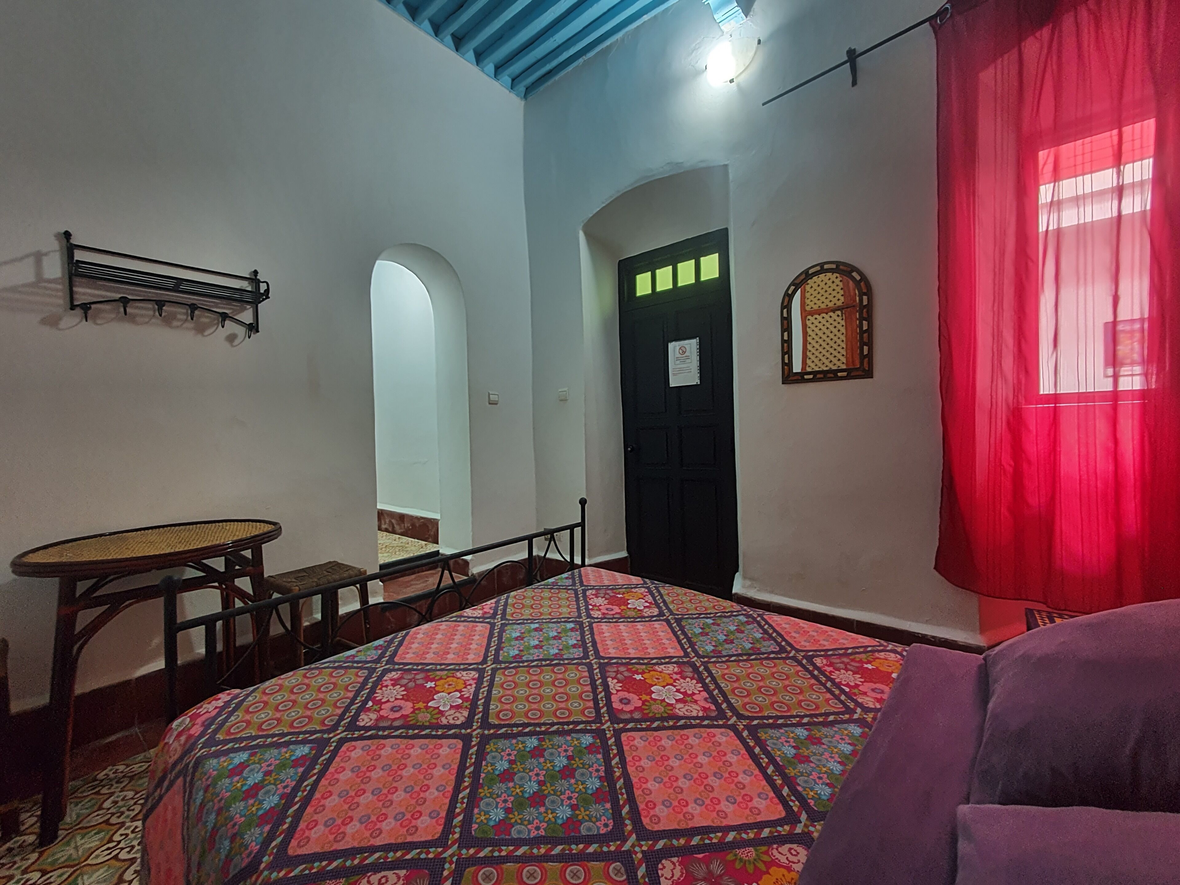 Una habitación con cama, una mesa y una silla. Tiene una puerta con un letrero, una ventana con cortina y una lámpara fijada a la pared.