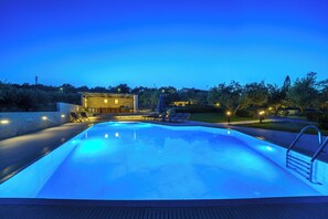 A swimming pool at dusk with lounge chairs and a building in the background. - Villas Lefkothea (Rethymno)