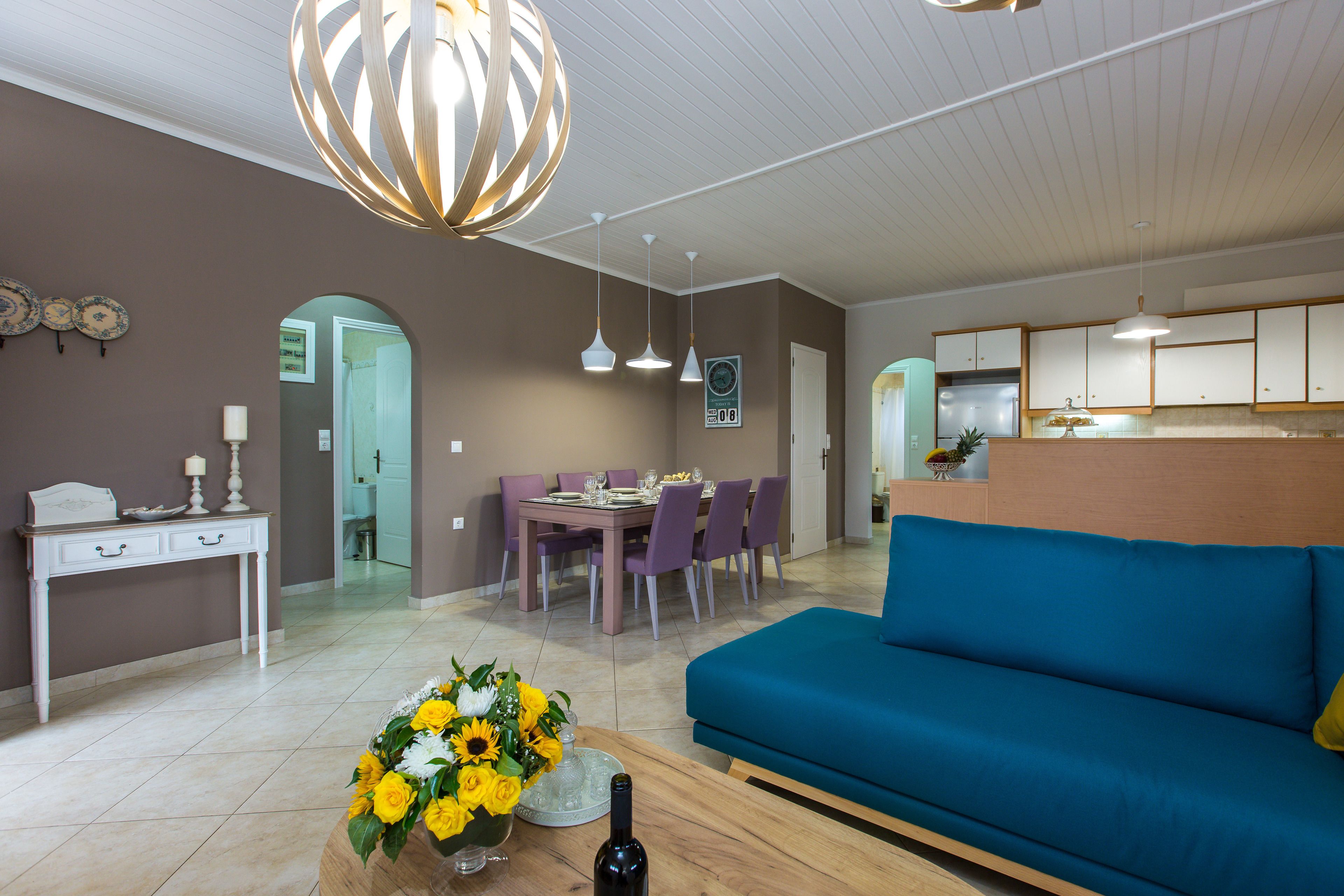 A modern living room with a blue sofa, a dining area with purple chairs, and a kitchen with white cabinets.