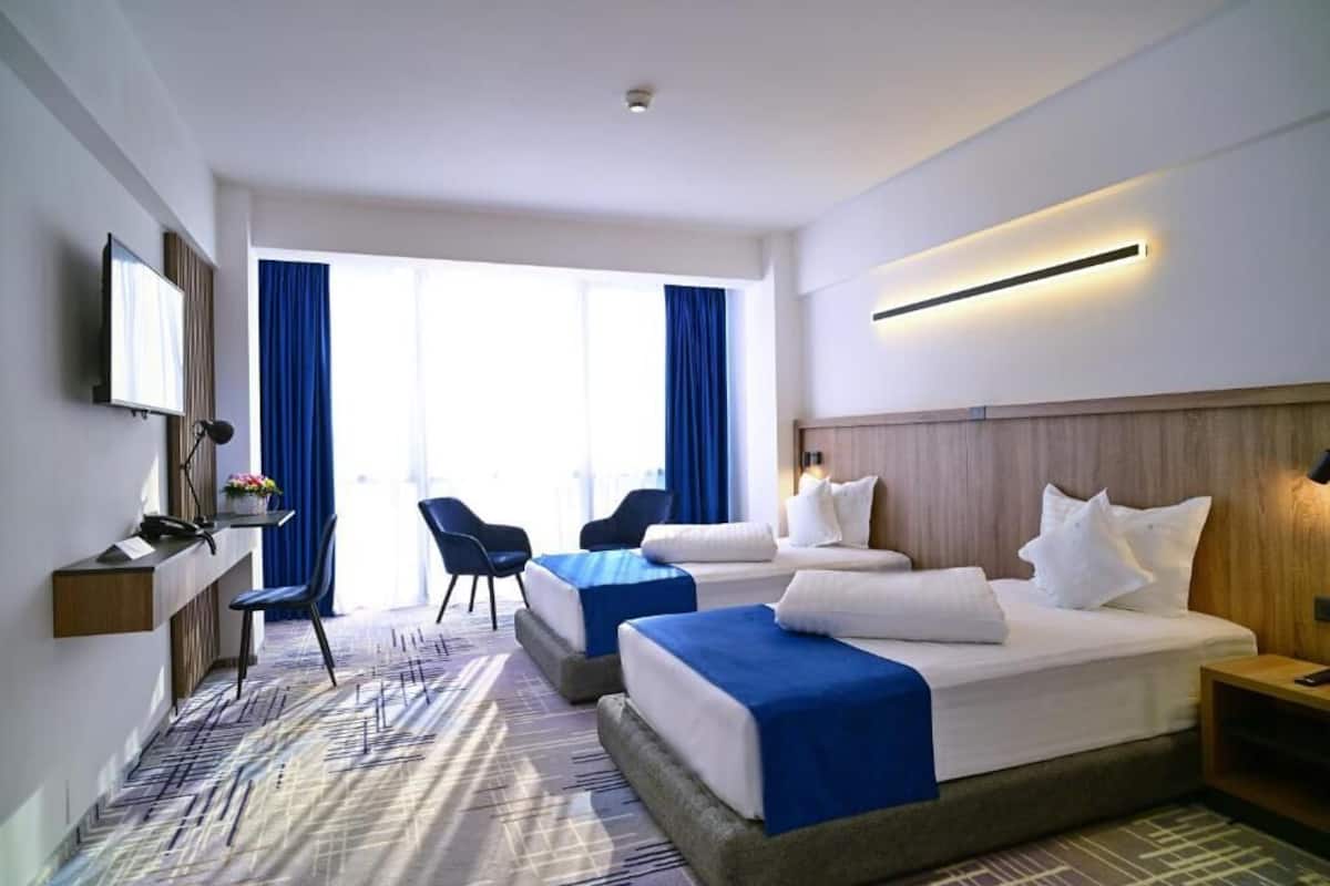 Habitación de hotel con dos camas, un escritorio, una silla y un ventanal con cortinas.