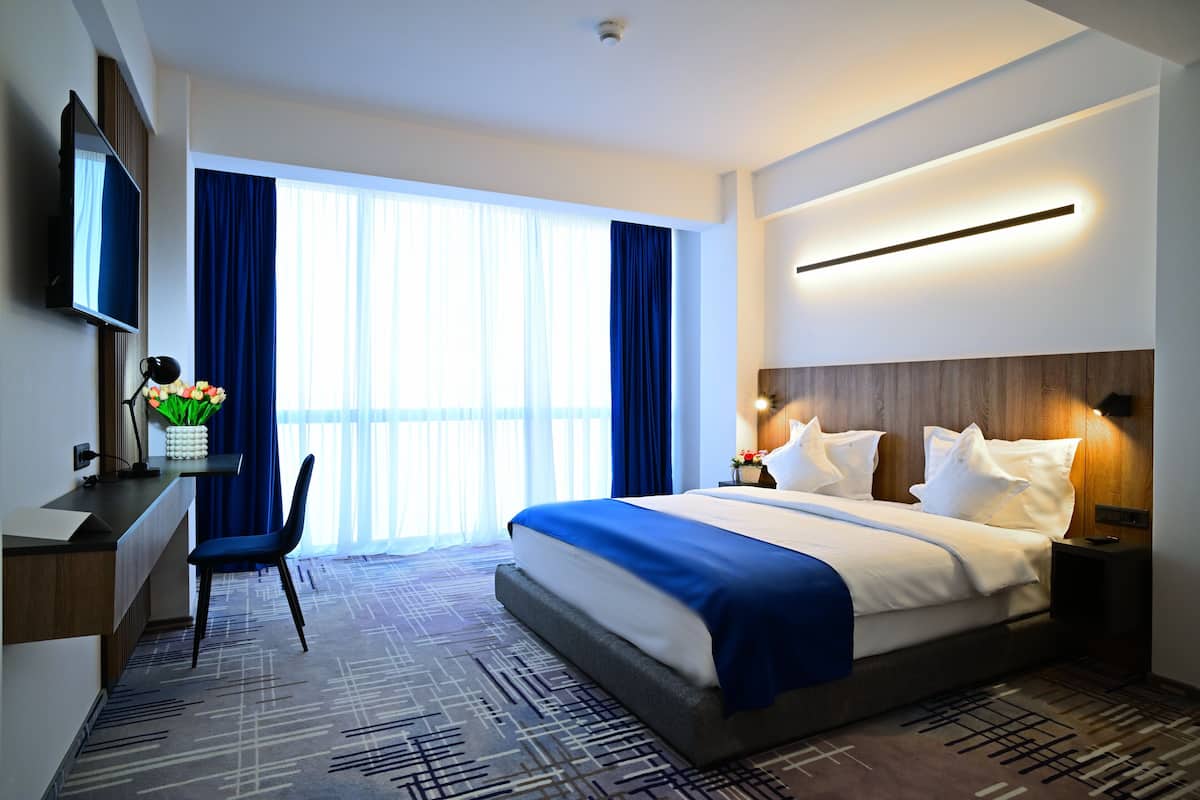 Una habitación de hotel con una cama grande, un escritorio con una silla, un televisor y un ventanal con cortinas.