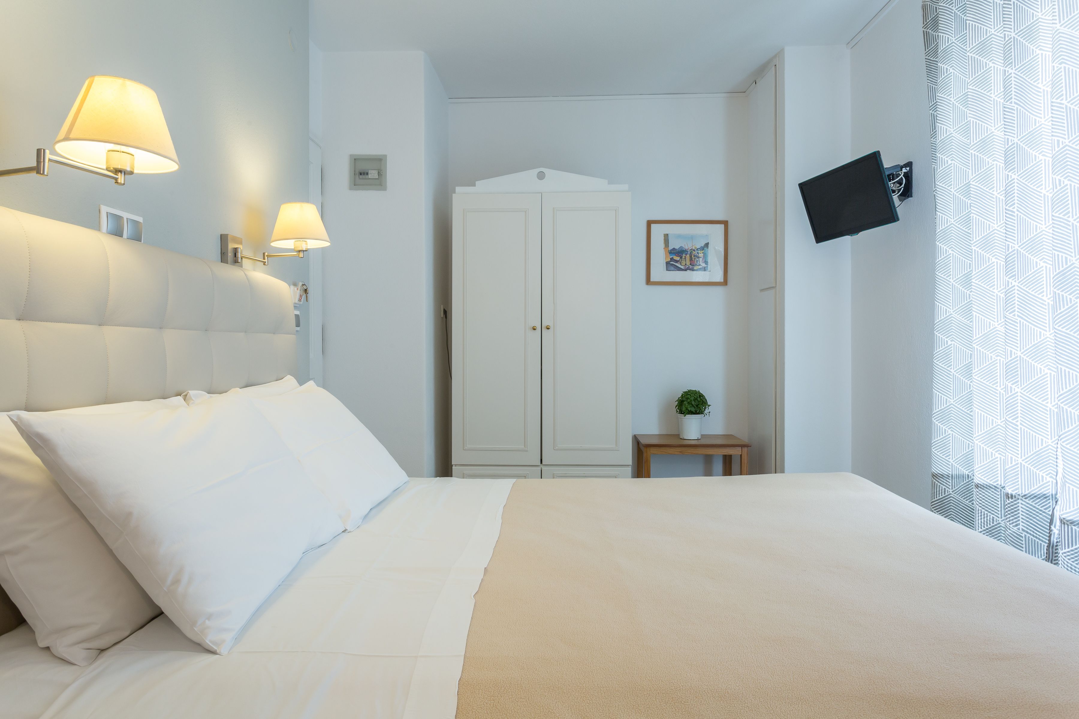 A hotel room with a bed, two wall-mounted lamps, a wardrobe, a small table with a plant, and a TV mounted on the wall.