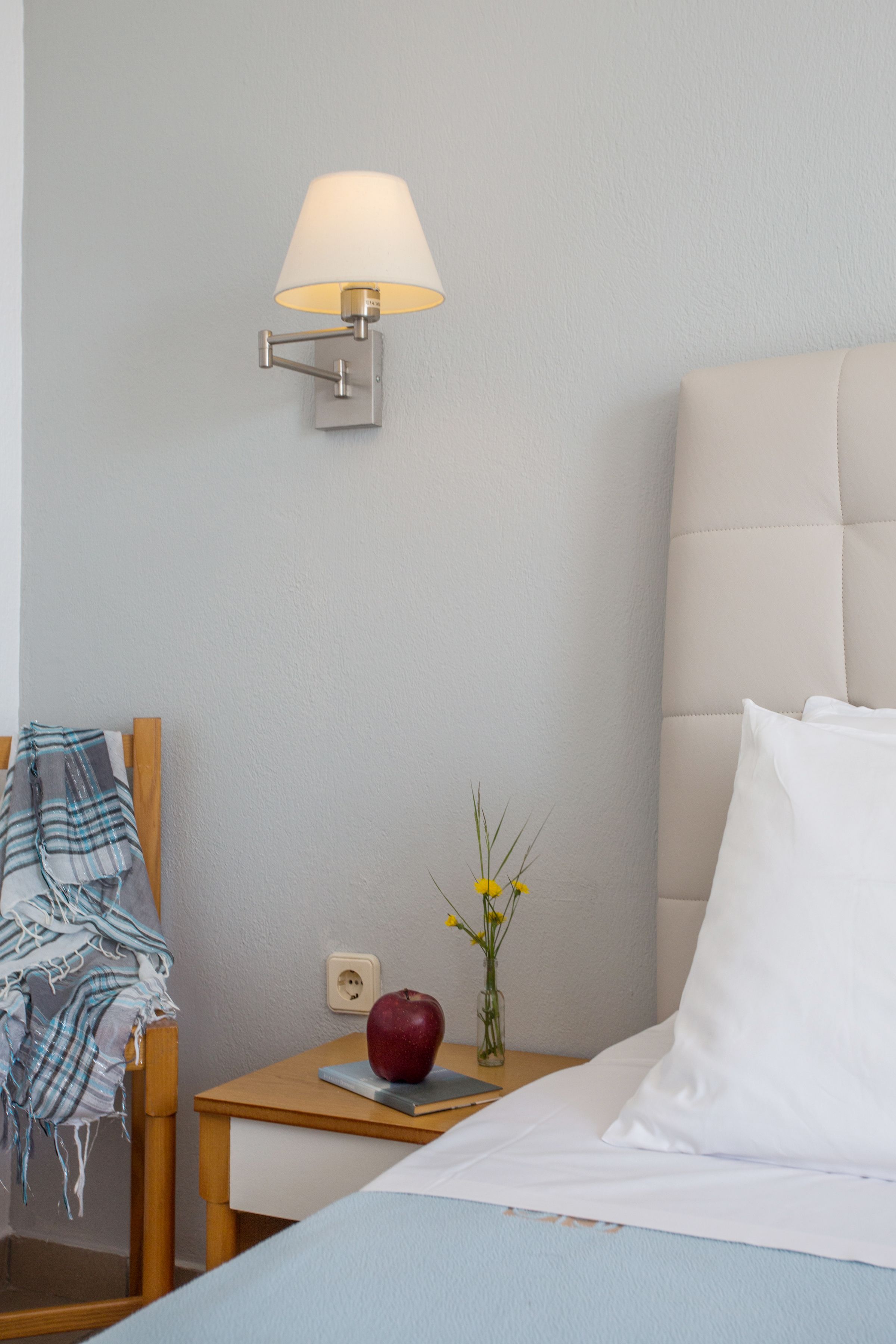 A neatly made bed with white pillows, a bedside lamp, a small wooden nightstand with a red apple and a vase of yellow flowers, and a chair with a plaid blanket.