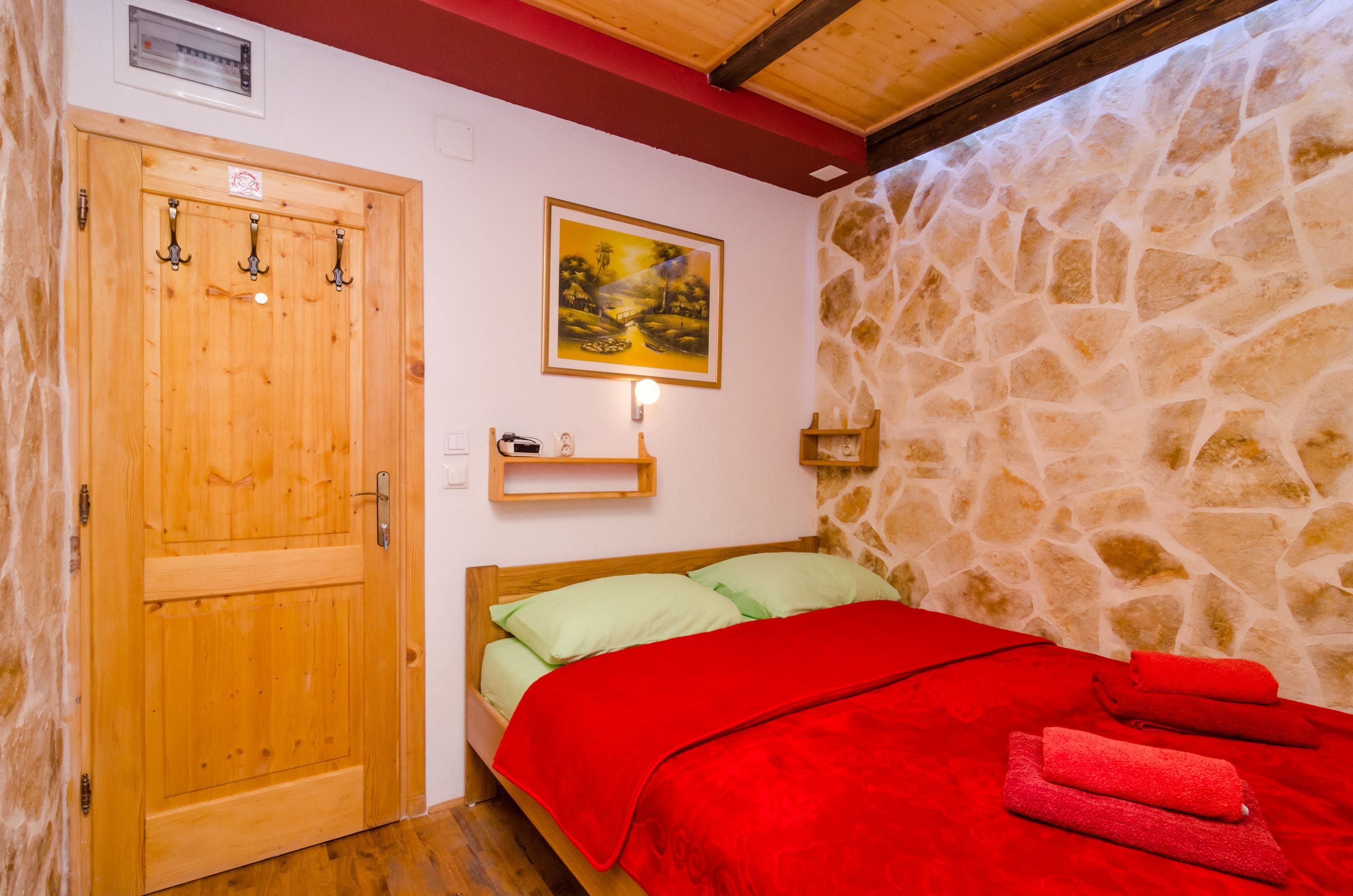 A room with a wooden door, a bed with green and red bedding, a stone wall, and a painting on the wall.