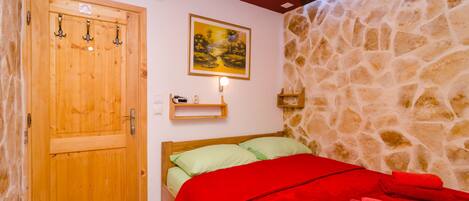 A room with a wooden door, a bed with green and red bedding, a stone wall, and a painting on the wall.