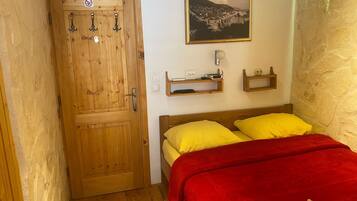 A room with a wooden door, a bed with yellow and red bedding, a wall-mounted shelf with a lamp, and a framed picture.