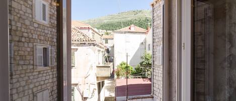 Visão pela janela de um edifício com paredes de pedra, telhado de telhas vermelhas e escadaria que leva a um andar superior.