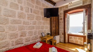 A room with stone walls, a wooden door leading to a balcony, a bed with red bedding, a small table with a picture frame, and a TV mounted on the wall. - Eddie´s Sea View Rooms (Dubrovnik)