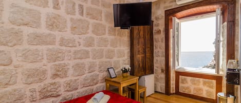 A room with stone walls, a wooden door leading to a balcony, a bed with red bedding, a small table with a picture frame, and a TV mounted on the wall.