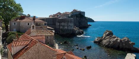 A coastal town with stone buildings, a fortified structure on a cliff, and clear blue sea.