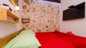 Una habitación con pared de piedra, una cama con ropa de cama roja y verde, un estante de madera, un reloj de pared y un televisor de pantalla plana.