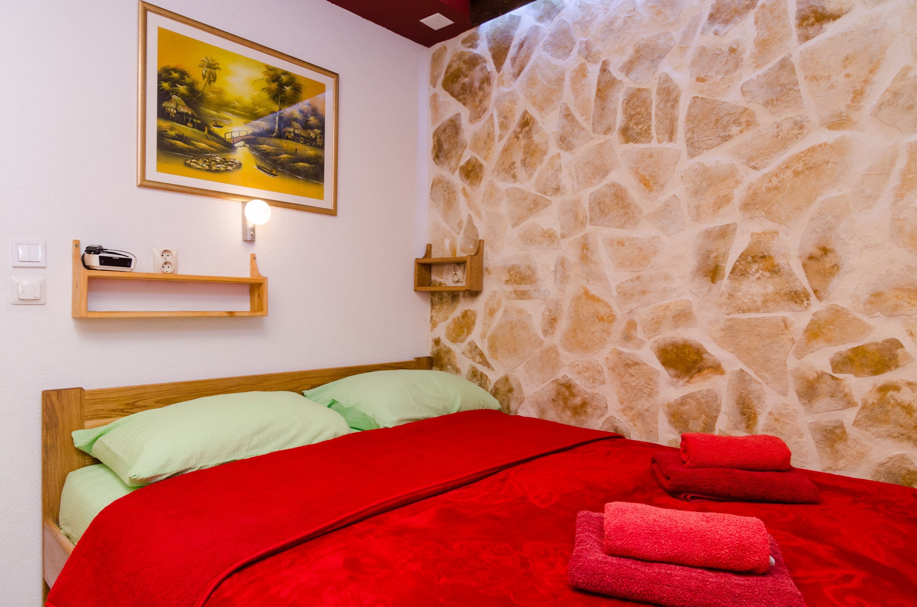 A bedroom with a stone wall, a bed with red bedding, a painting on the wall, and a shelf with a radio.