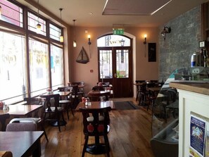 An interior with wooden tables and chairs, a counter with a menu board, and large windows allowing natural light.