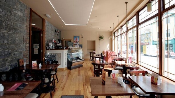 Interno di un ristorante con tavoli e sedie in legno, una parete in pietra e ampie finestre.