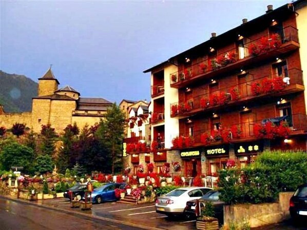A hotel with multiple balconies adorned with red flowers, a stone building in the background, and parked cars in front.
