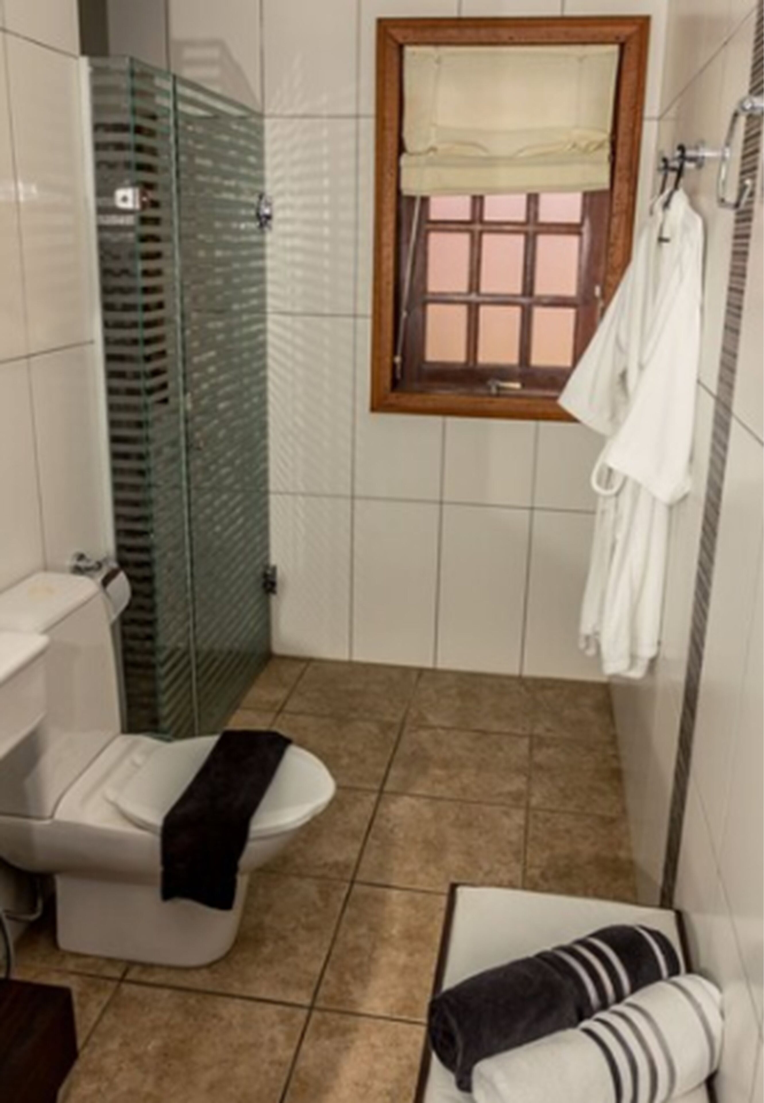 A small bathroom with a toilet, a glass shower enclosure, a window with a wooden frame, and a towel rack with white towels.