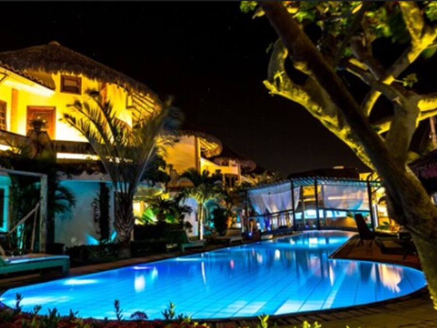 Une vue nocturne d’un hôtel avec une piscine, des palmiers et un bâtiment au toit de chaume.