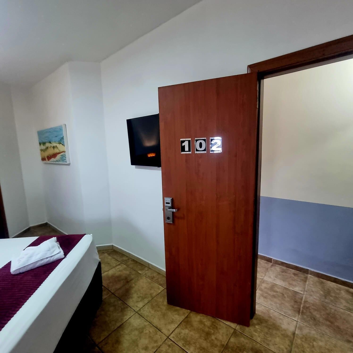 Een hotelkamer met een bed, een aan de muur bevestigde televisie en een deur met het nummer 102.