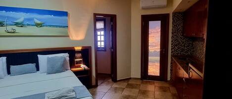 A hotel room with a bed, a painting of sailboats on the wall, a kitchen area, and a view through an open door.