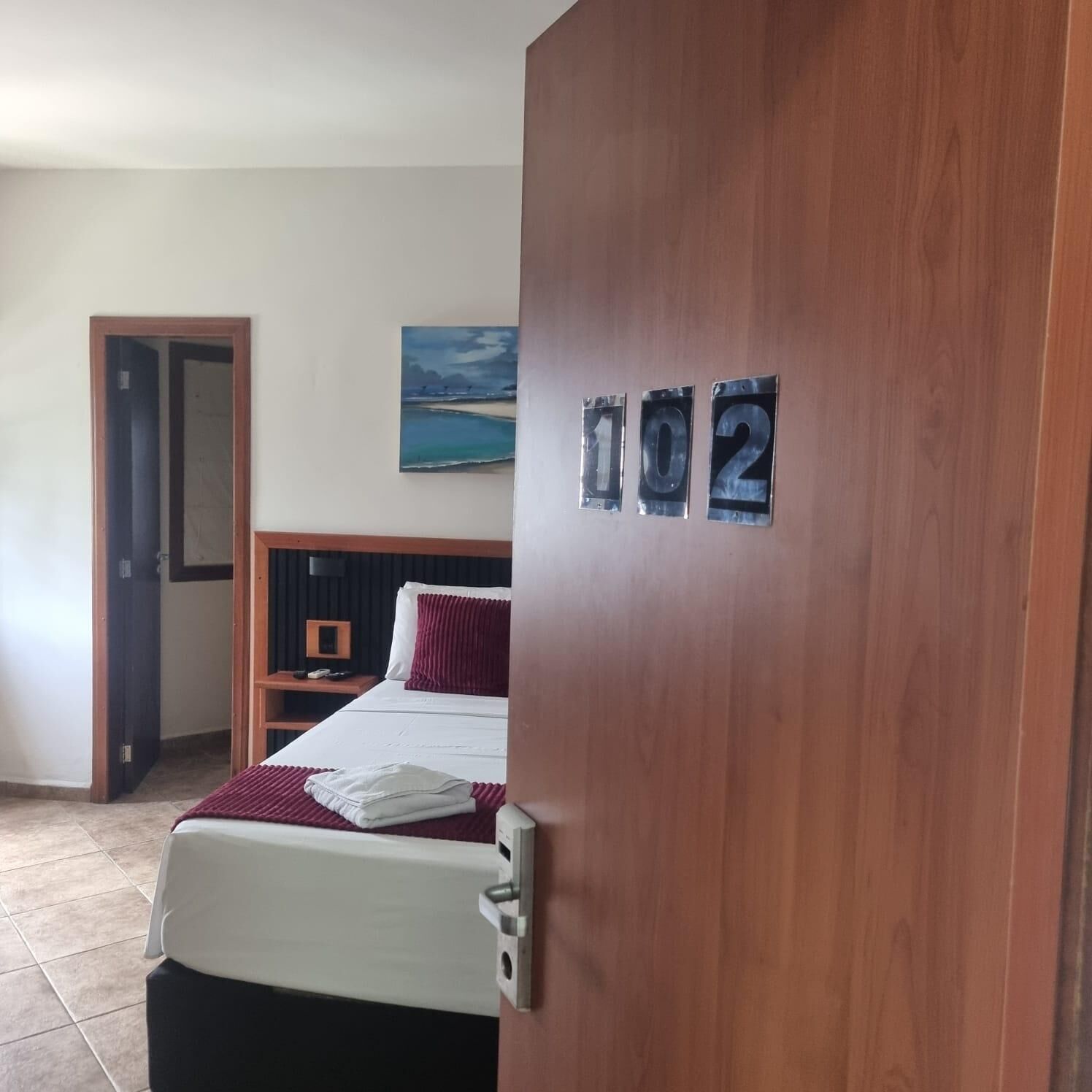 A hotel room with a bed, a nightstand, a painting, and a door with numbers 102 on it.