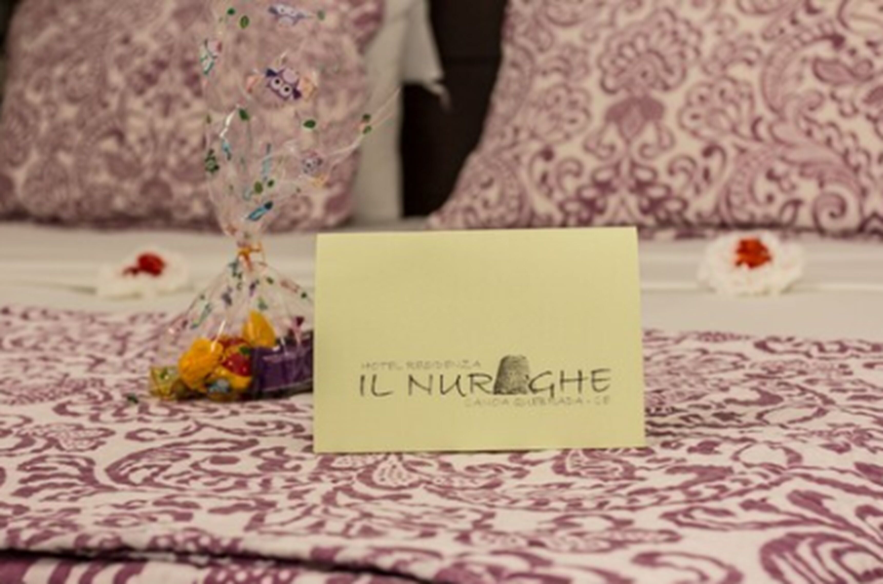Una habitación de hotel con una cama cubierta por una colcha estampada, una mesita con una tarjeta amarilla y una guirnalda decorativa de adornos coloridos.