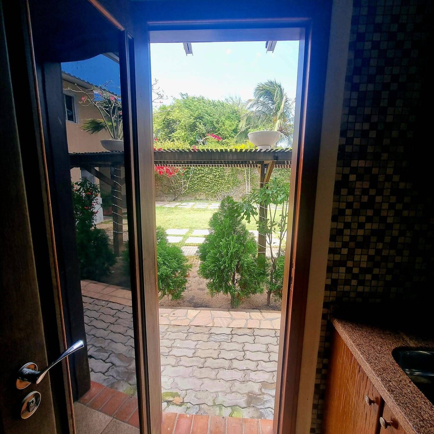 Vista a través de un ventanal hacia un patio con suelo de baldosas, una pérgola y diversas plantas.