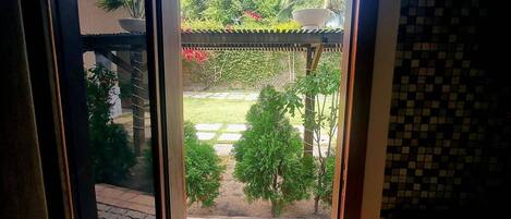 Vista a través de un ventanal hacia un patio con suelo de baldosas, una pérgola y diversas plantas.
