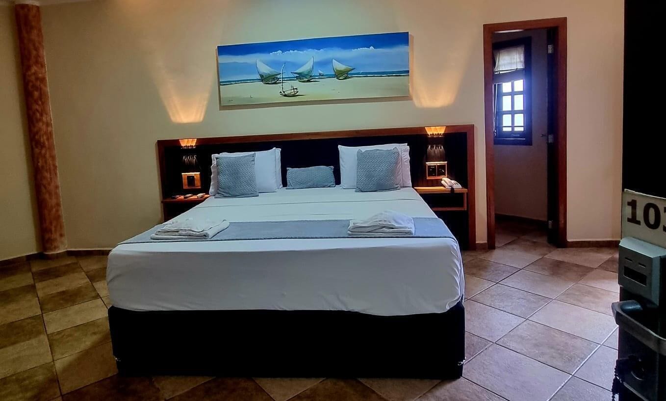 Ein Hotelzimmer mit einem großen Bett, einem Segelboot-Gemälde an der Wand, einem Nachttisch mit einer Lampe und einer Tür zu einem weiteren Raum.