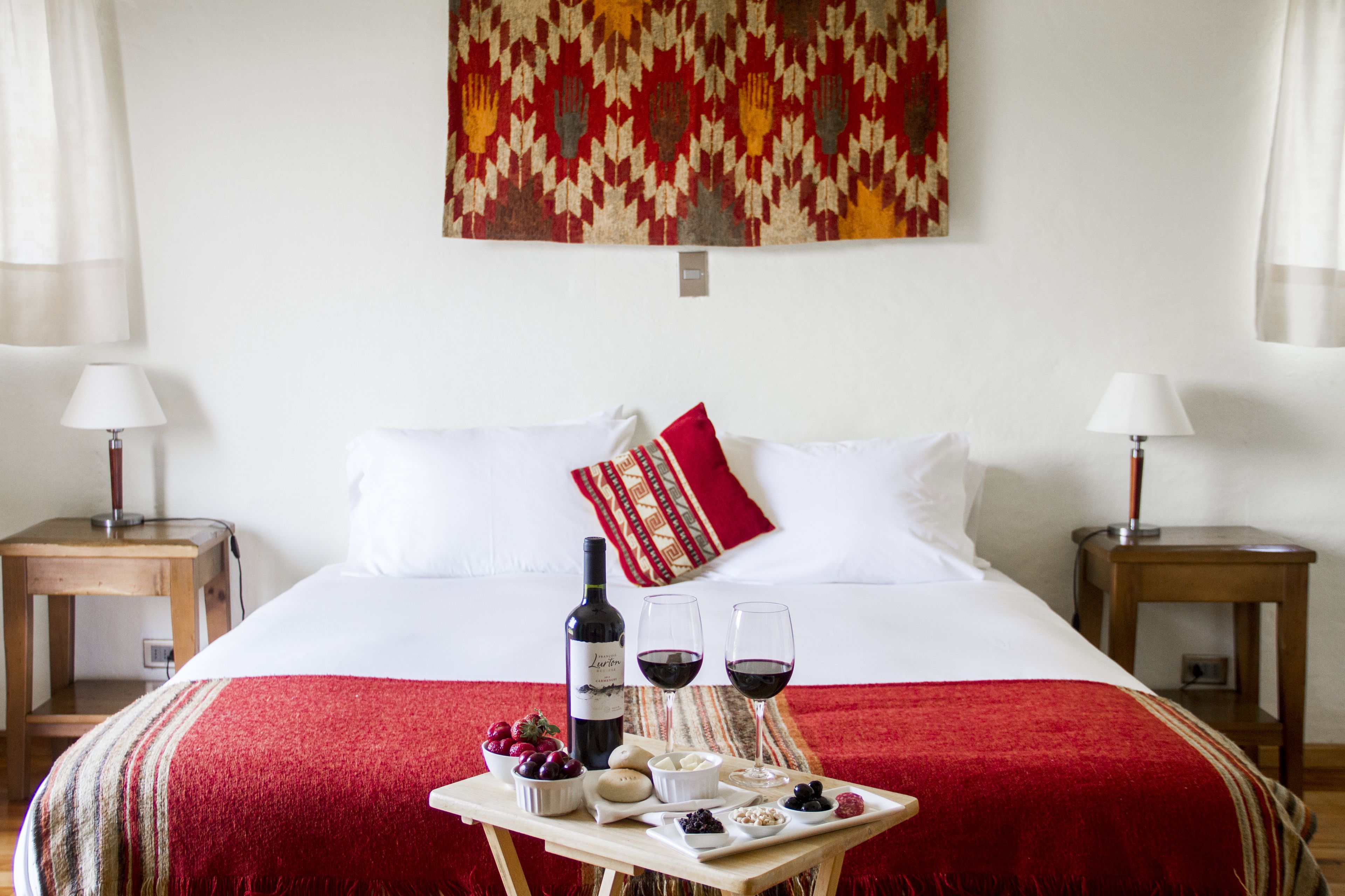 Una habitación de hotel con una cama, una mesa con vino y aperitivos, y un tapiz decorativo.