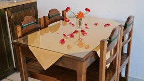 A dining table with a glass top and a beige table runner. There are four wooden chairs around the table. A vase with flowers and rose petals is on the table.