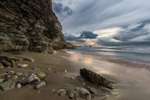 A rocky beach with a large cliff, calm sea, and a sunset in the distance.