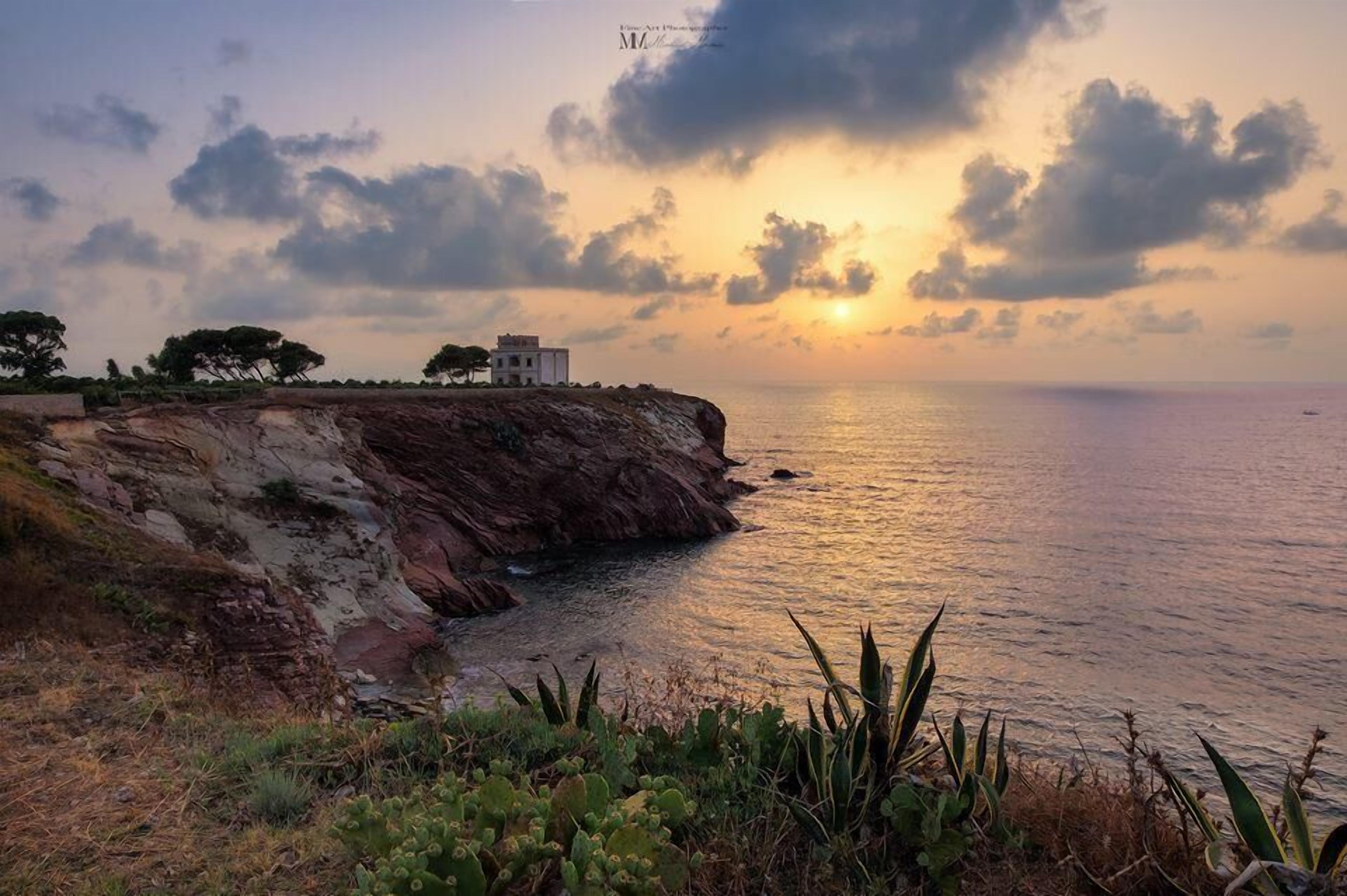 A coastal landscape with a cliff, vegetation, and a building overlooking the sea.