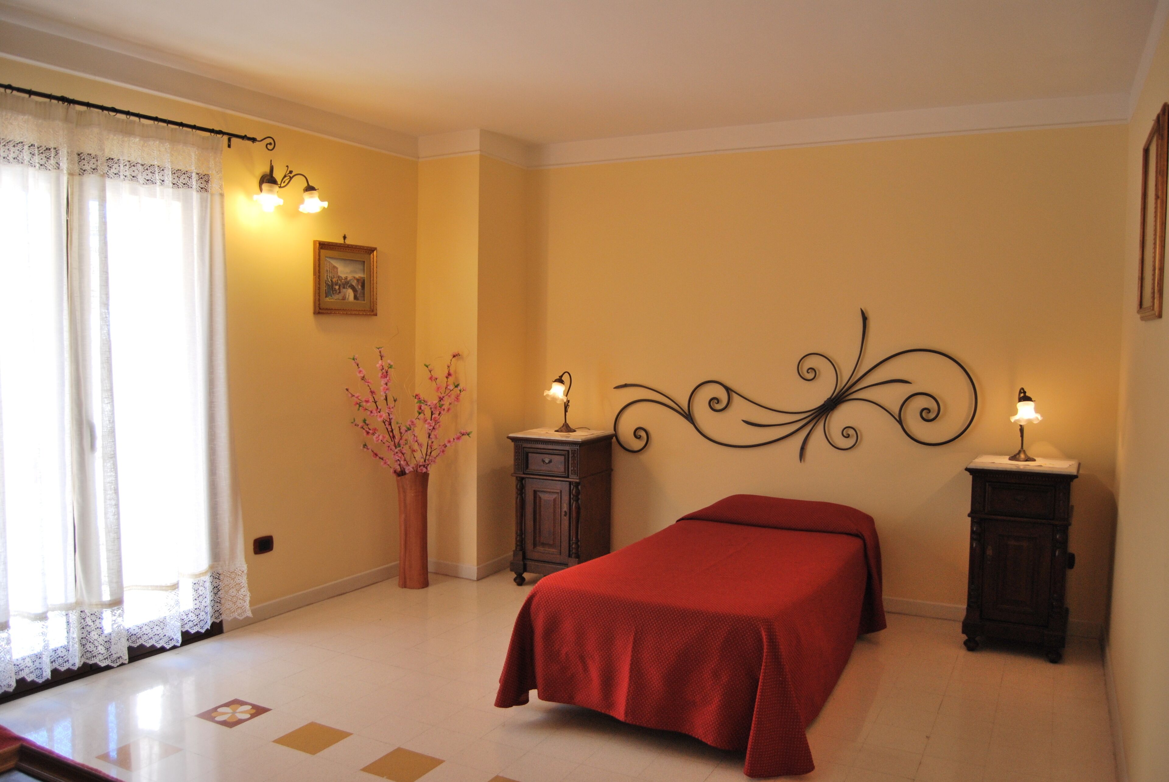 Un dormitorio con cama, dos mesitas de noche, lámpara de pared, un cuadro y un jarrón con flores.