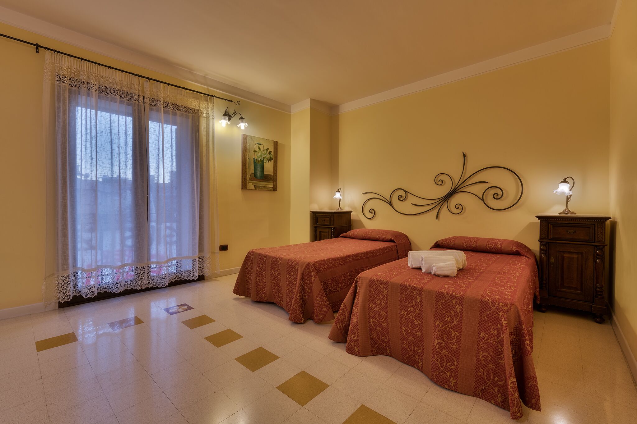 Une chambre d’hôtel avec deux lits, une grande fenêtre avec des rideaux légers, un élément décoratif mural et une petite table avec une lampe.