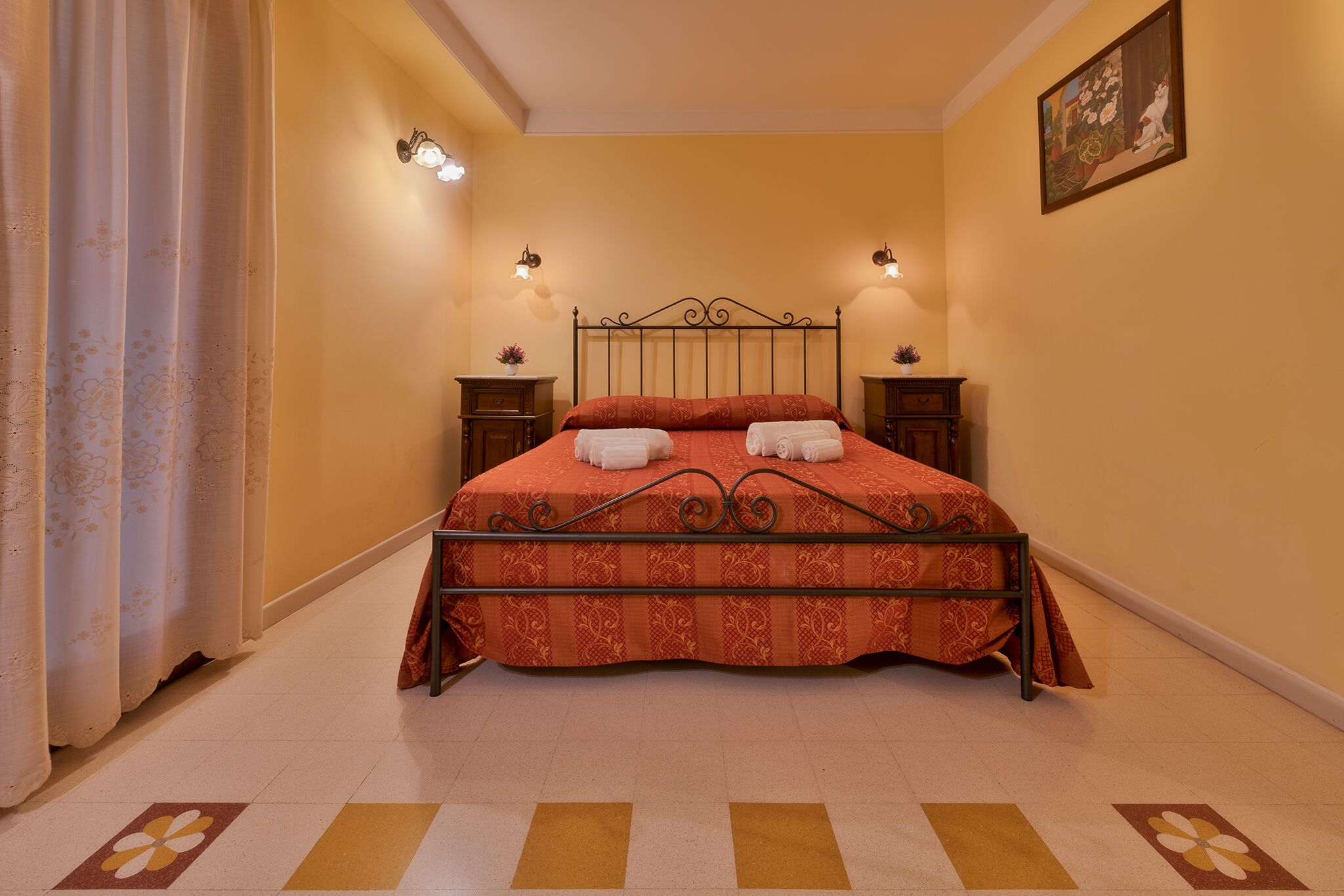 A neatly made bed with a patterned bedspread and two towels, two bedside tables with lamps, and a painting on the wall.