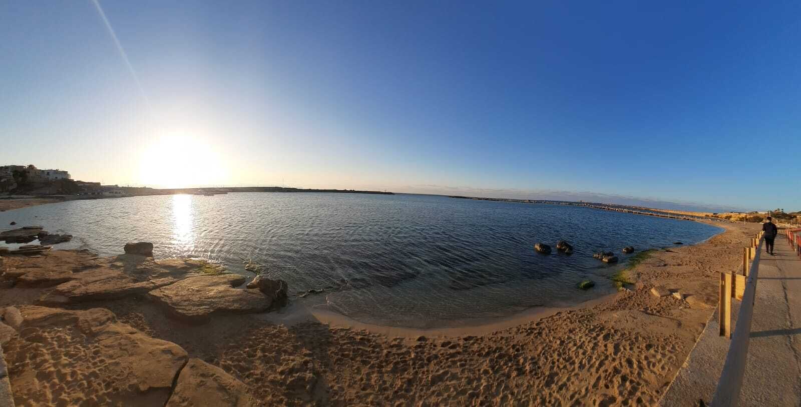 A beach with a clear sky, a person walking on the path, and rocks near the water.