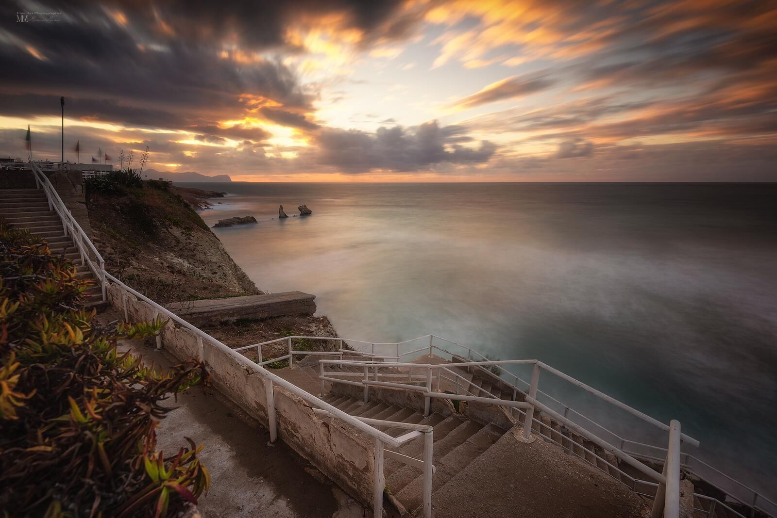 A coastal staircase leading to a viewpoint overlooking the sea, with a sunset in the background.