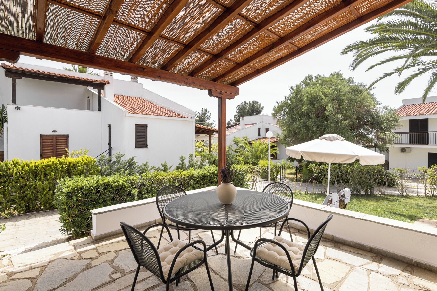 A covered outdoor seating area with a round table and chairs, surrounded by greenery and buildings.