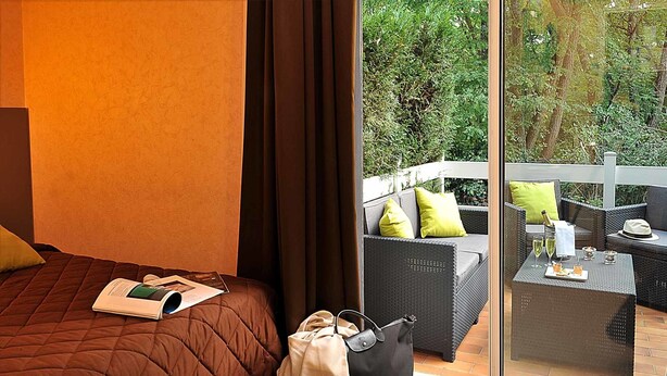A hotel room with a bed, a window with a view of greenery, and a balcony with outdoor furniture. - Hôtel Le Prieuré (Saint-Marcel)