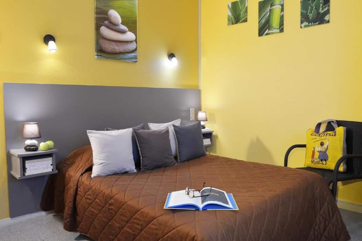 A bedroom with a brown bedspread, two pillows, a bedside lamp, a nightstand with books and a lamp, and a wall-mounted light fixture.